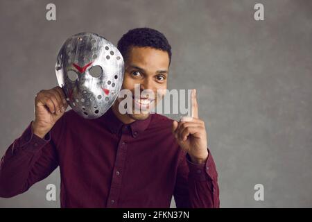 Felice bel giovane uomo nero toglie la sua brutta maschera spaventosa, punta il dito verso l'alto e sorride Foto Stock