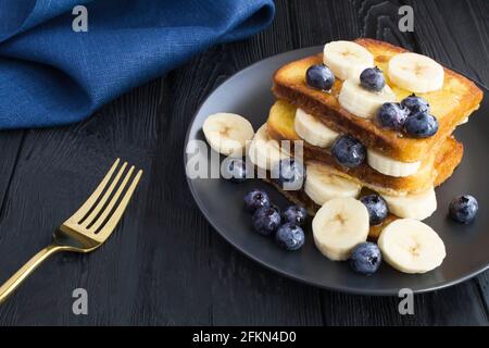 Toast francesi con mirtilli, banana e miele nella piastra nera su fondo di legno scuro. Primo piano. Foto Stock