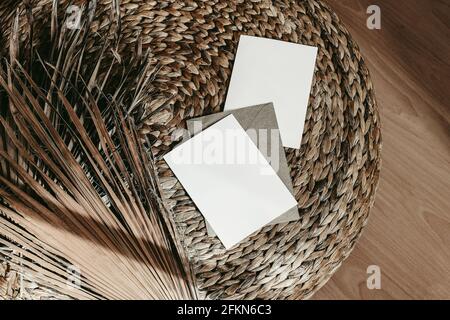 Mockup di cancelleria estiva tropicale. Biglietti d'auguri vuoti, buste artigianali e foglie di palma asciutte. Pouff di paglia di rattan su pavimento di legno. Boho moderno Foto Stock