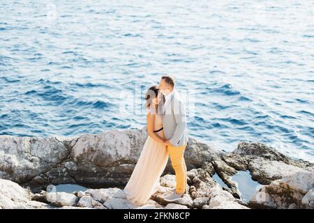 Lo sposo tiene le mani della sposa mentre si trova sulle rocce sopra il mare blu. Vista dall'alto Foto Stock