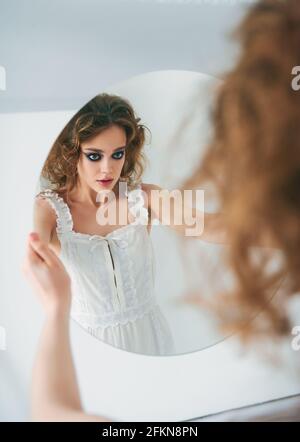 Ritratto di una ragazza piuttosto strana. Bella donna dispari in abito bianco guardando nello specchio Foto Stock