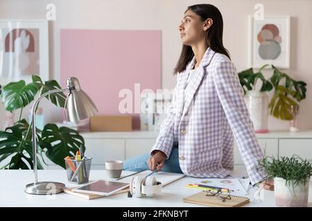 Giovane donna sicura con un abbigliamento casual elegante che sorride mentre si è in piedi vicino al suo posto di lavoro Foto Stock