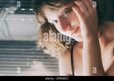 giovane donna con occhi blu e capelli corti all'interno della sua casa, seduta accanto alla cieca chiusa dove il suo corpo si riflette nella finestra Foto Stock