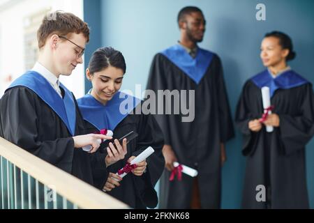 Diversi gruppi di giovani che indossano abiti da laurea e utilizzano smartphone all'interno dell'università, spazio di copia Foto Stock