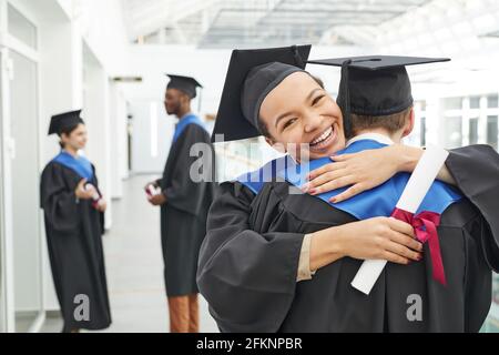 Ritratto di felice afroamericana ragazza con abito graduato hugging amico e sorridente a macchina fotografica, copia spazio Foto Stock