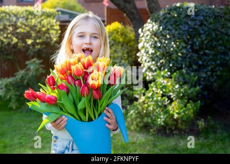 Carino adorabile sorridente ragazza senza denti con un enorme mazzo di tulipani nel annaffiatoio. Raccolta fiori per bouquet regalo in giardino. Madre Foto Stock