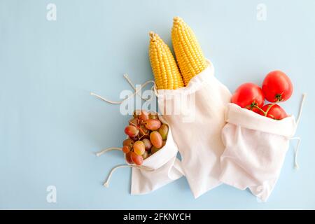 Due pannocchie di mais, uva e rametto di pomodori ciliegini in tre sacchetti riutilizzabili di cotone eco-friendly con lacci su sfondo azzurro. Zero rifiuti c Foto Stock