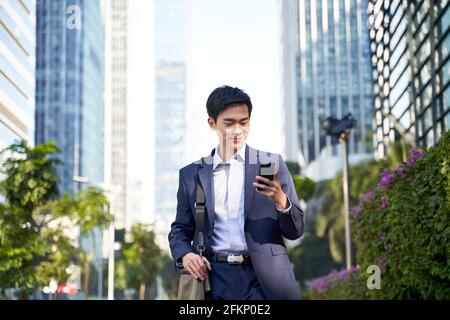 giovane uomo d'affari asiatico che guarda i messaggi sul cellulare mentre cammina nella strada nel centro della città moderna Foto Stock