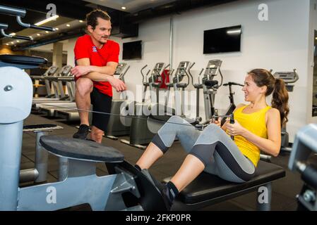 Giovane atleta caucasica nel moderno centro fitness che fa esercizio sulla palestra mentre chiacchiera con un allenatore personale. Foto Stock