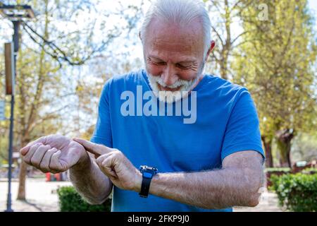 Uomo anziano che controlla il polso dopo l'addestramento nel parco. L'anziano sportivo che controlla la frequenza cardiaca monitorando il suo smartwatch sul polso. Foto Stock