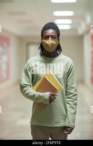 Ritratto verticale di un giovane afroamericano che indossa una maschera a scuola corridoio e guardando la macchina fotografica Foto Stock