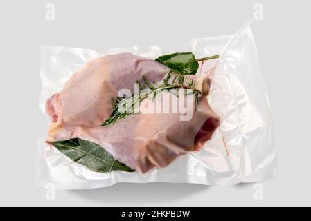 Cosce di pollo confezionate sottovuoto sigillate per cottura a sous vide con rosmarino, foglia di alloro e salvia, isolate su sfondo grigio chiaro Foto Stock