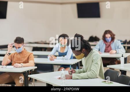 Vista laterale ritratto di giovane afroamericano uomo che indossa maschera mentre si prende prova o esame a scuola con diversi gruppi di persone, spazio copia Foto Stock