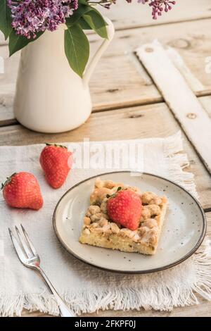 Pezzo di torta crumble di rabarbaro e fragole su un piatto su un vecchio tavolo di legno, decorato con fiori di lilla, verticale Foto Stock