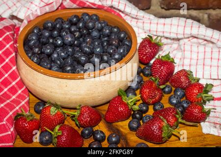 Una ciotola rustica in ceramica, riempita con mirtilli colorati Indigo e circondata da più mirtilli e fragole Foto Stock