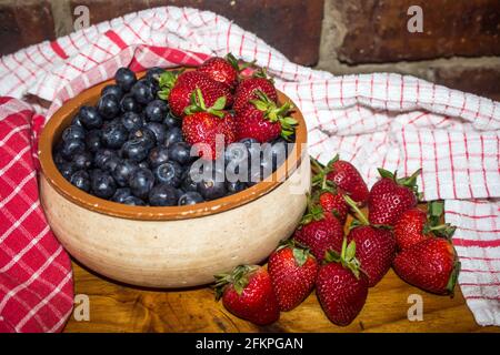 Una ciotola rustica in ceramica, piena di mirtilli, circondata da fragole e asciugamani rossi da cucina Foto Stock