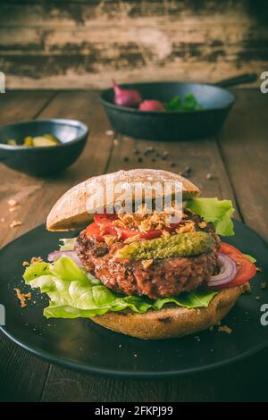 Hamburger vegetariano con insalata, cetriolo, pomodori e cipolle arrostite su un piatto nero su tavolo di legno, foto verticale stock Foto Stock