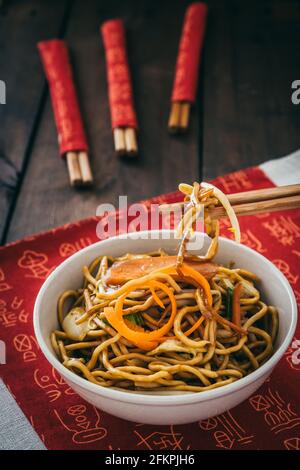 Tagliatelle vegetariane cinesi in una ciotola bianca su tappetino cinese posto su tavolo di legno, foto verticale stock Foto Stock