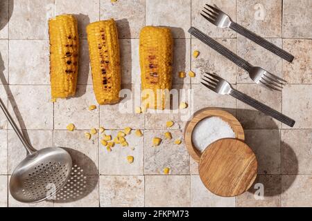 Deliziose pannocchie di mais alla griglia con tre forchette, cantina di sale, cucchiaio a taglio su storie di pietra toniche di terra vista dall'alto Foto Stock