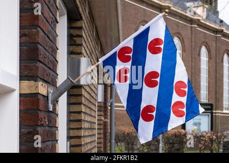Bandiera frisone tricolore, bandiera provinciale della provincia della Frisia, pende da un flagpole contro un muro a Koudum, Paesi Bassi. Chiesa sullo sfondo Foto Stock