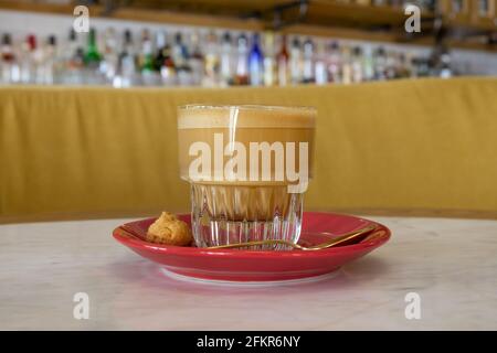 Un'elegante tazza di vetro con caffè bianco piatto servito con un piattino rosso, cucchiaio dorato e piccoli biscotti. Caffè nella caffetteria. Messa a fuoco selettiva, bac sfocato Foto Stock