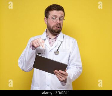 Il medico sorpreso tiene una tavoletta in mano e punta eccitatamente il dito sullo schermo. Sfondo giallo. Foto Stock