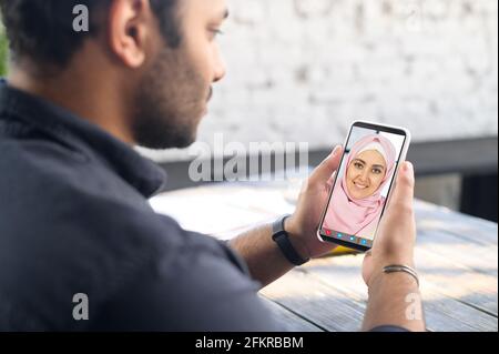 Indietro view multirazziale uomo medio-orientale utilizzando l'app per cellulare per video riunione con un amico o un dipendente che indossa hijab, ragazzo tiene smartphone, parlando online con la donna islamica. Videochiamata Foto Stock