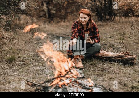 Viaggiatrice donna in campeggio nella foresta e rilassarsi vicino al falò dopo una giornata molto dura. Concetto di trekking, avventura e vacanza stagionale. Foto Stock