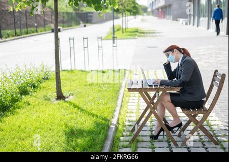 Una ragazza stanca con gli occhiali lavora a distanza su un computer all'aperto. Una donna d'affari in una maschera sta scrivendo su un computer portatile mentre si siede su una terrazza estiva Foto Stock