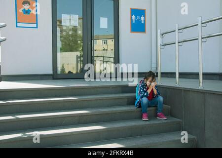 Ragazza piccola triste in maschera protettiva seduta sul scale della scuola Foto Stock