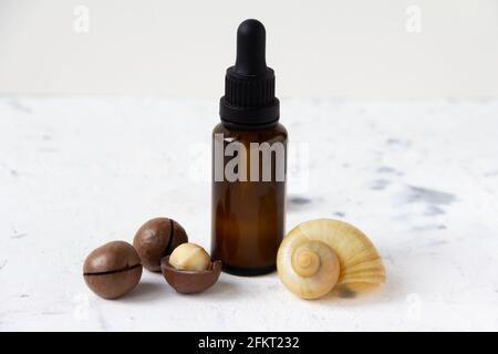 Flacone cosmetico contagocce in vetro scuro con noci di macadamia e guscio di lumaca su sfondo bianco. Foto Stock