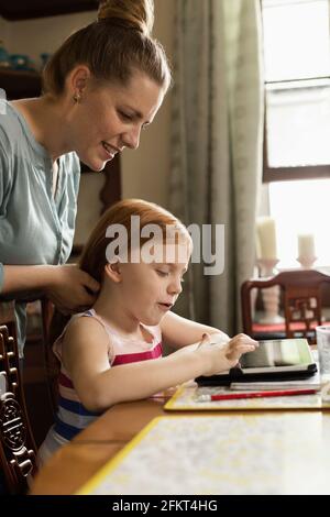 Ragazza con tavoletta digitale al tavolo da pranzo mentre la madre lo styling i capelli Foto Stock