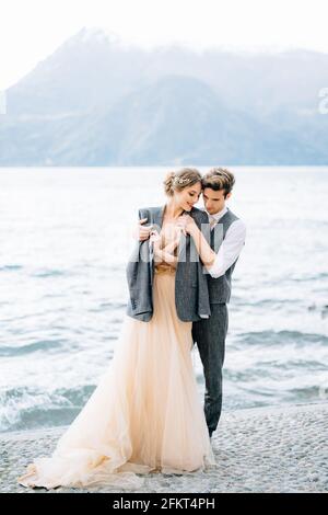 Lo sposo gettò una giacca sulle spalle della sposa che abbracciava Lei sul lungofiume del lago di Como vicino all'acqua Foto Stock