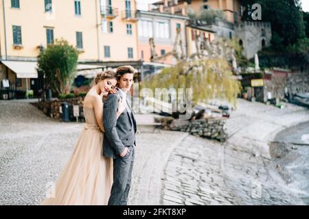 Sposa abbracca lo sposo da dietro, appoggiando la testa sulla spalla, in piedi sul terrapieno del lago di Como sullo sfondo di vecchie case Foto Stock