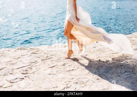 Le gambe della sposa in sandali dai tacchi alti e con una gonna che si gonna in fluttering, la sposa cammina lungo il molo, da vicino Foto Stock