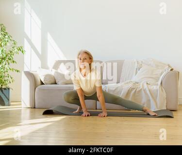 Belle donne sane e mature degli anni '60 nello sport indossano esercizi di stretching sul tappetino yoga nel soggiorno a casa. Anziane donne in pensione si riscaldano al mattino. Sport e stile di vita sano in pensione Foto Stock