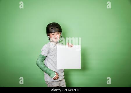 giovane scolaro fresco con braccio rotto e gesso verde con elmetto nero che tiene la lavagna bianca davanti allo sfondo verde Foto Stock