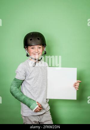 giovane scolaro fresco con braccio rotto e gesso verde con elmetto nero che tiene la lavagna bianca davanti allo sfondo verde Foto Stock