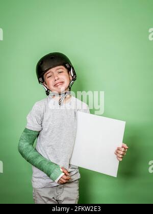 giovane scolaro fresco con braccio rotto e gesso verde con elmetto nero che tiene la lavagna bianca davanti allo sfondo verde Foto Stock