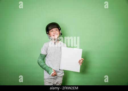 giovane scolaro fresco con braccio rotto e gesso verde con elmetto nero che tiene la lavagna bianca davanti allo sfondo verde Foto Stock