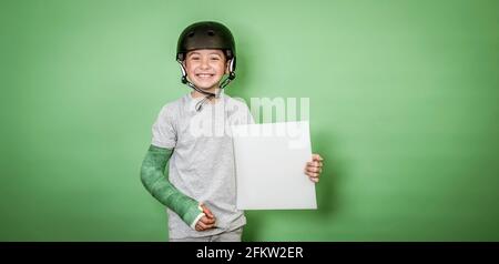 giovane scolaro fresco con braccio rotto e gesso verde con elmetto nero che tiene la lavagna bianca davanti allo sfondo verde Foto Stock