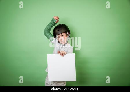 giovane scolaro fresco con braccio rotto e gesso verde con elmetto nero che tiene la lavagna bianca davanti allo sfondo verde Foto Stock