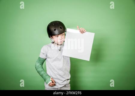 giovane scolaro fresco con braccio rotto e gesso verde con elmetto nero che tiene la lavagna bianca davanti allo sfondo verde Foto Stock