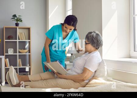 Infermiera di cura domestica che parla con la donna anziana distesa che sta sedendo su letto e libro di lettura Foto Stock