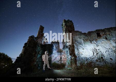 Vista laterale dello spaceman in speciale tuta spaziale bianca a piedi vicino alle antiche rovine e guardando le stelle di notte. Concetto di ricerca antiche rovine. Foto Stock