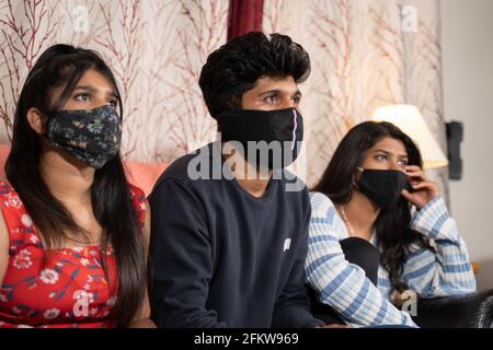 Focus su giovane uomo, fratelli con maschera medica di faccia occupato guardando film o web serie on-line o notizie covid mentre si siede sul divano durante blocco dovuto Foto Stock