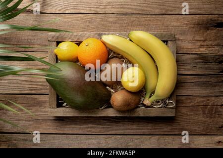 Frutta tropicale fresca in una scatola di consegna di legno su sfondo di legno. Arancione, banana, mango, kiwi e limone vista dall'alto piatto con spazio per la copia per som Foto Stock