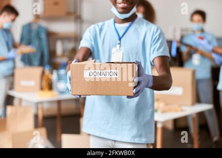 Primo piano di scatola di cartone per la donazione in mani di giovane volontario africano americano maschio in uniforme blu, maschera protettiva e guanti. Team Foto Stock