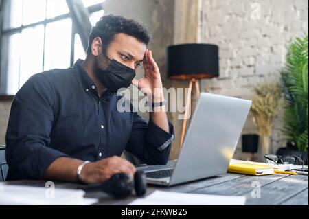 Stanco e preoccupato impiegato indù maschio indossando maschera medica protettiva fissando allo schermo del portatile, uomo misto-razza che lavora in ufficio durante le malattie virali si diffonde, proteggendosi con la maschera Foto Stock