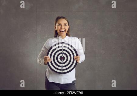 Giovane donna ridente forte che tiene il bersaglio della tavola di freccette in piedi sopra parete dello studio Foto Stock